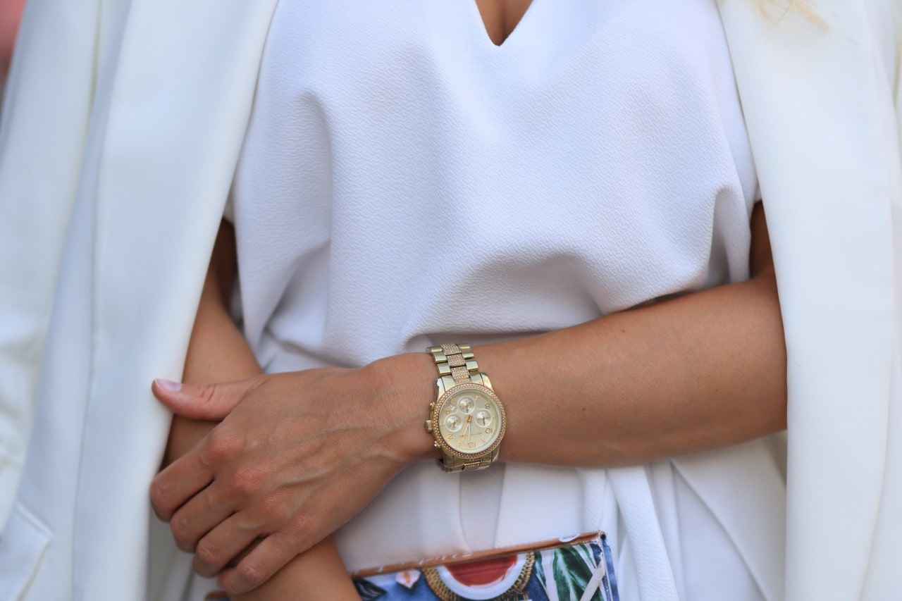 Close up of woman wearing all white with a gold watch on her wrist, holding a colorful clutch in the other hand