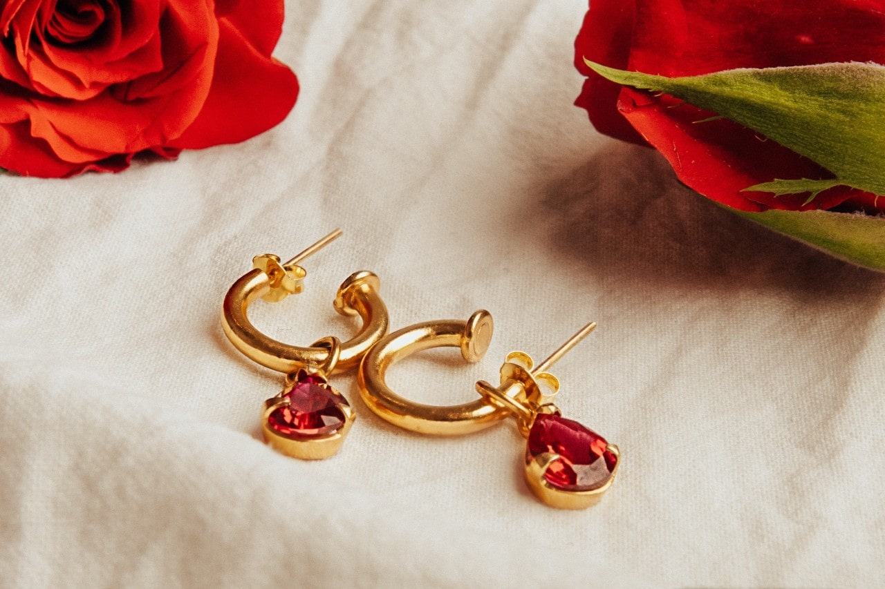 A pair of gold hoop and red gemstone earrings on a white cloth next to red roses