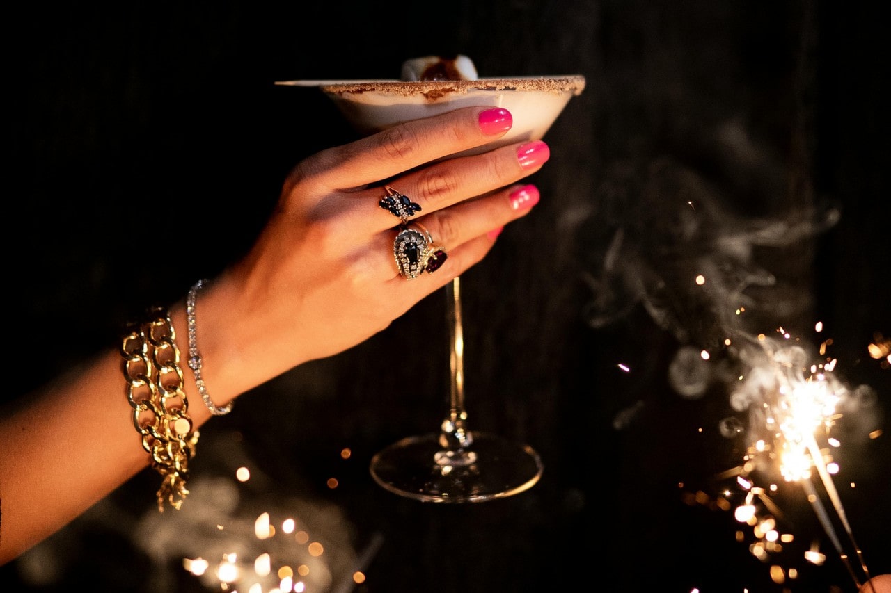 A hand with pink nails and adorned with elegant gold bracelets and rings, holds a cocktail glass. Bright sparklers illuminate the dark, festive background.