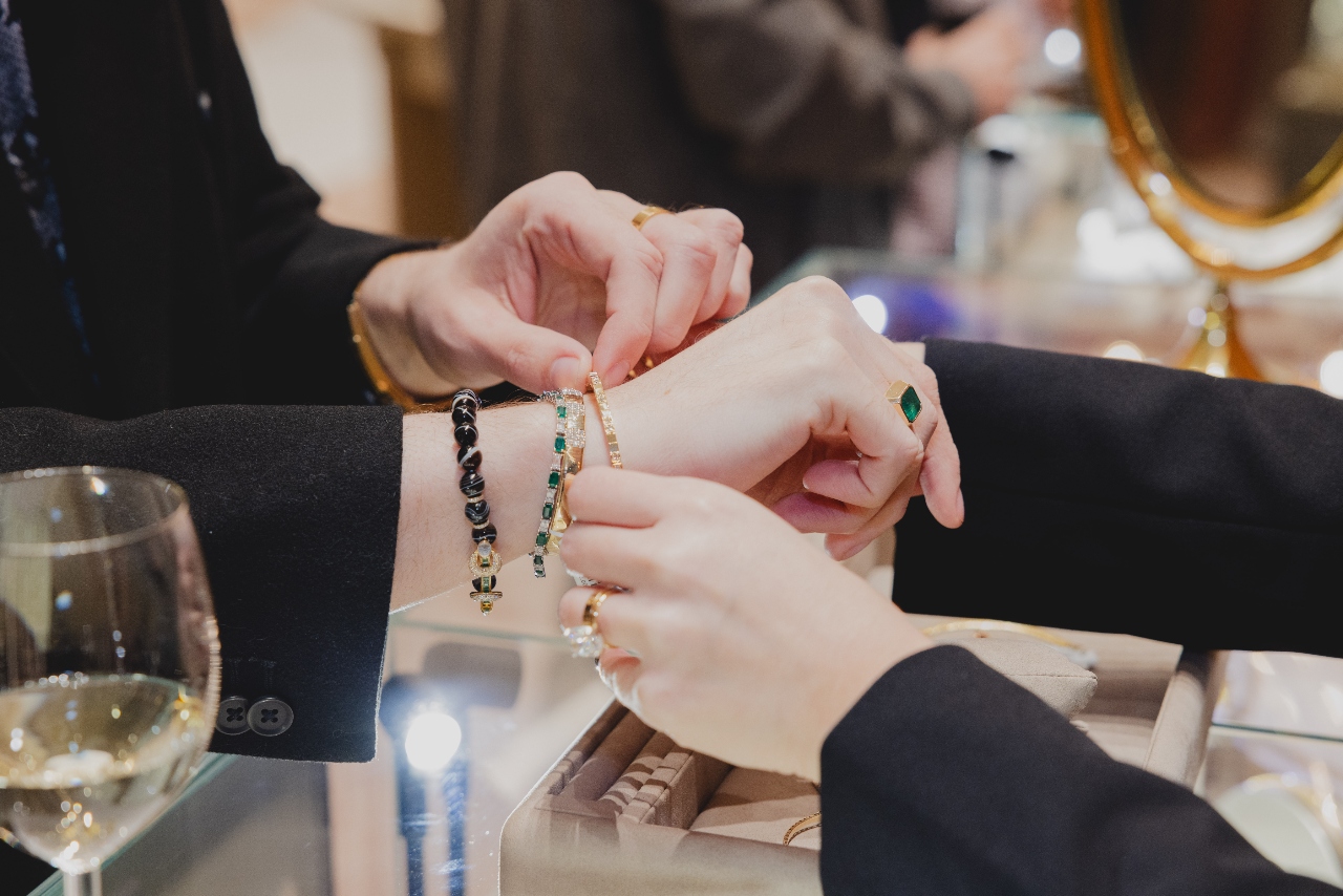 Person having a yellow gold and diamond bracelet adjusted on their wrist over a jeweler’s counter while they also wear black beaded bracelet, an emerald and diamond bracelet, and an emerald ring.