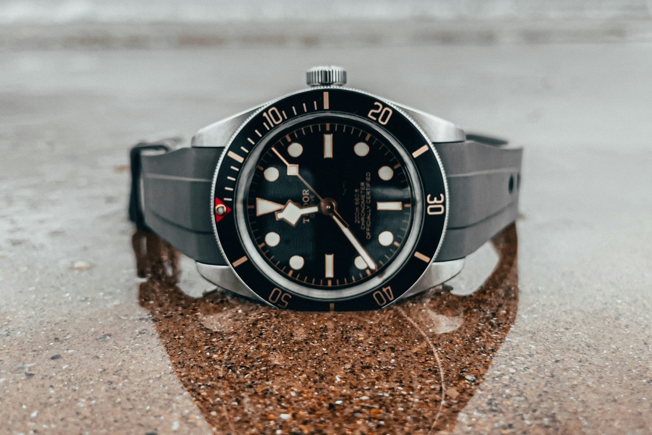 A close-up of a sturdy yet elegant TUDOR watch displayed on its side atop a reflective graphite surface.