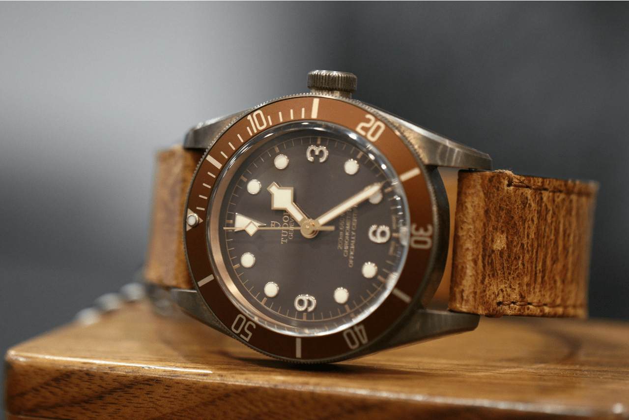 A close-up of a vintage-inspired TUDOR watch with a brown leather strap displayed on its side atop a wooden surface.