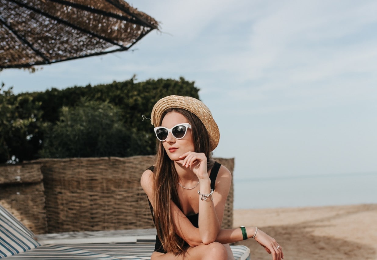 a woman sitting on a beach in a hat and sunglasses and jewelry