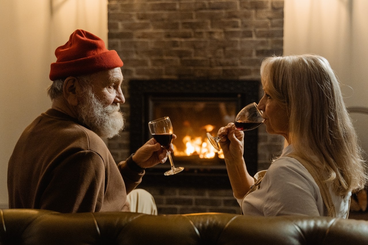 An older couple sips red wine by the fireplace in celebration.