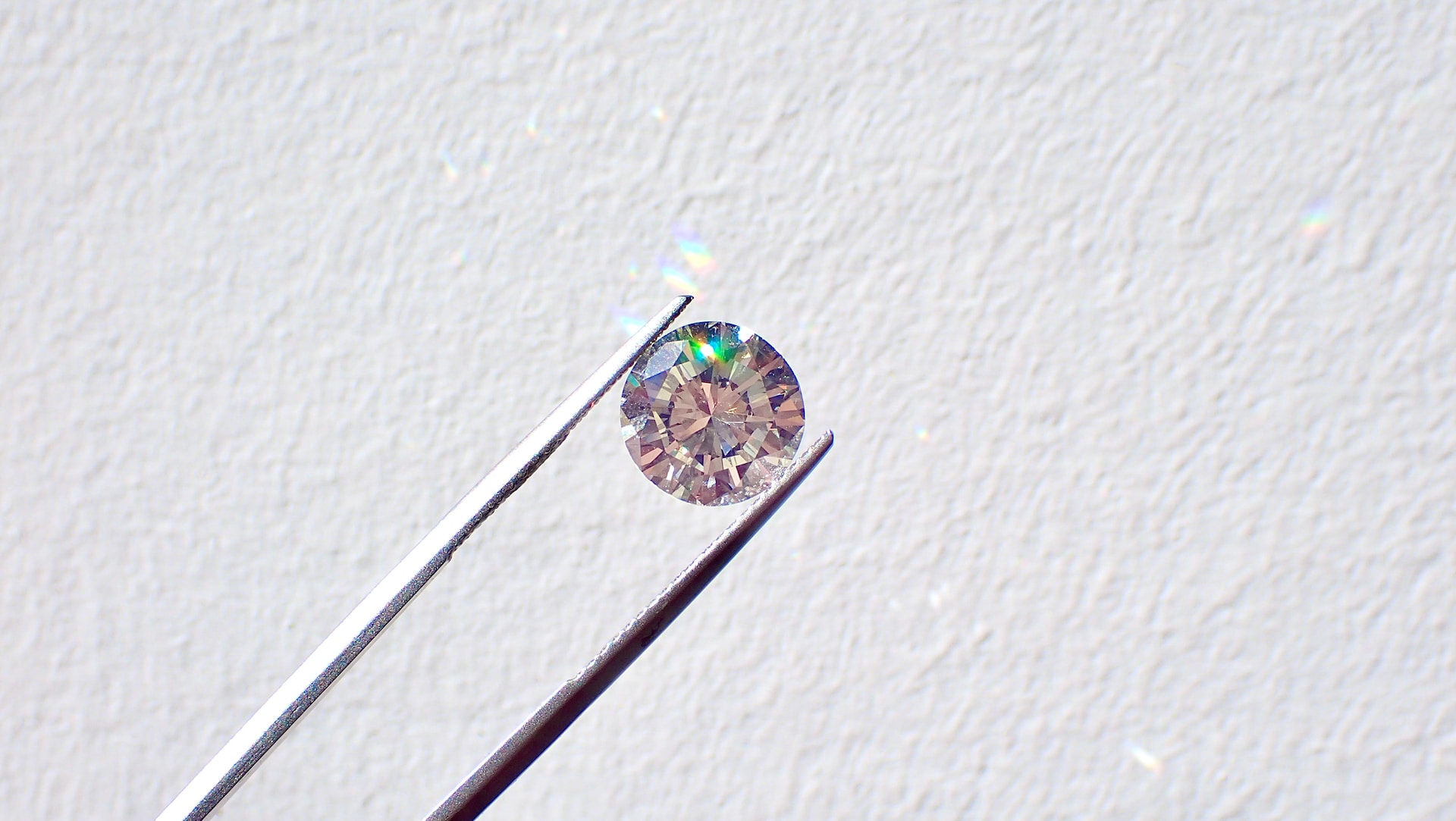 A pair of jewelry pliers holds up a round-cut diamond against a white wall.