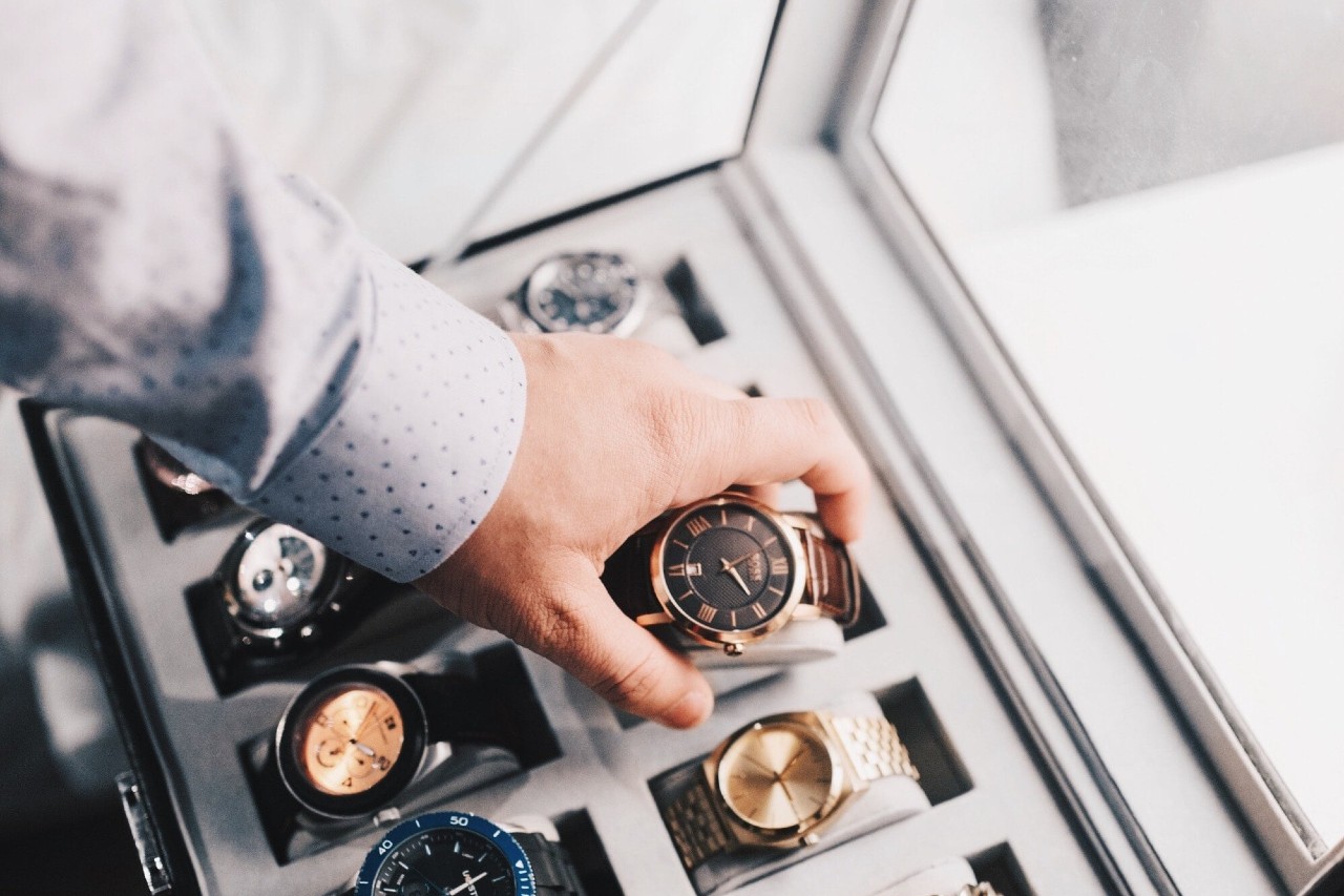 A photo of a man in a patterned dress shirt selecting a luxury wristwatch from a display case filled with multiple luxury timepieces.