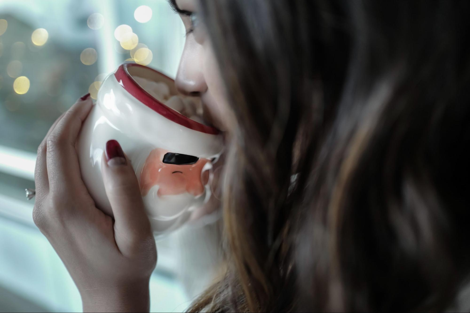 An engaged woman sips hot chocolate out of a Santa mug while watching the lights.