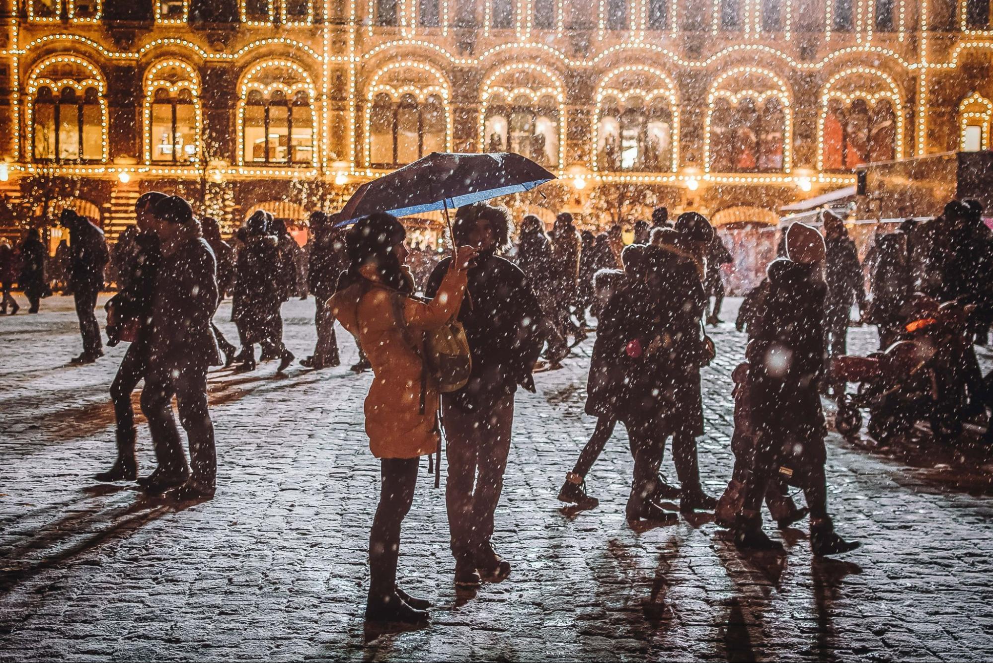 A couple under an umbrella walk through a lit plaza as the snow falls.