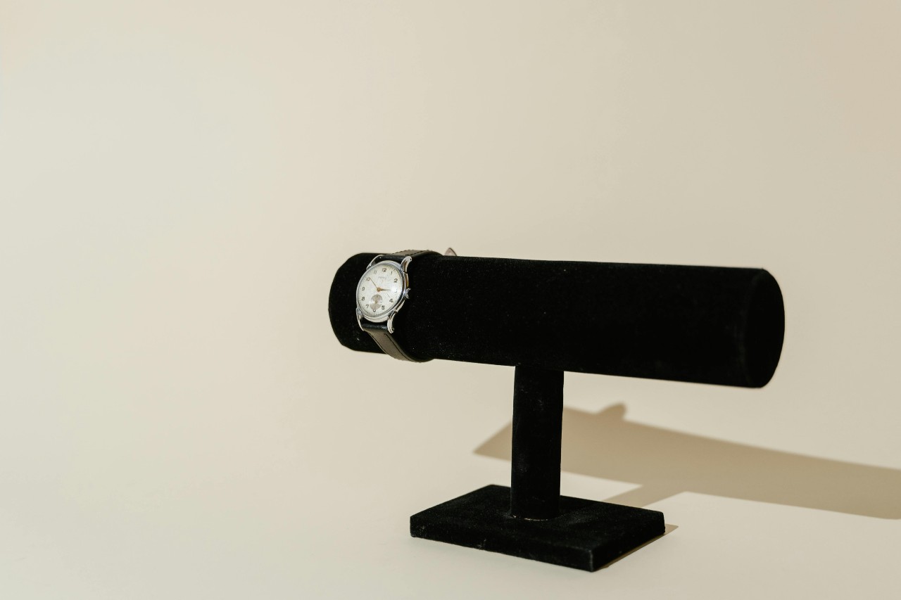 A white and black watch on a black watch stand in front of a beige backdrop.