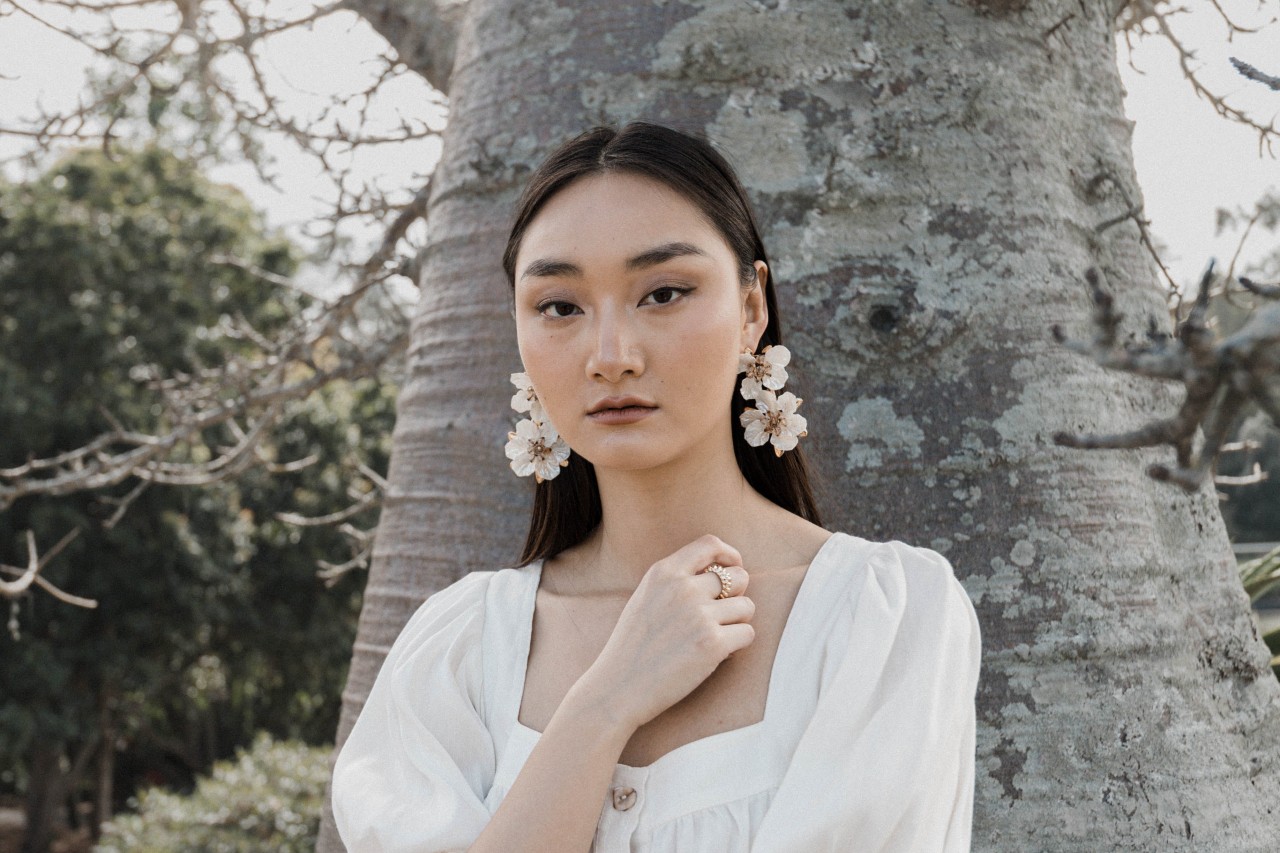 a woman standing in front of a tree wearing a white top and white floral earrings