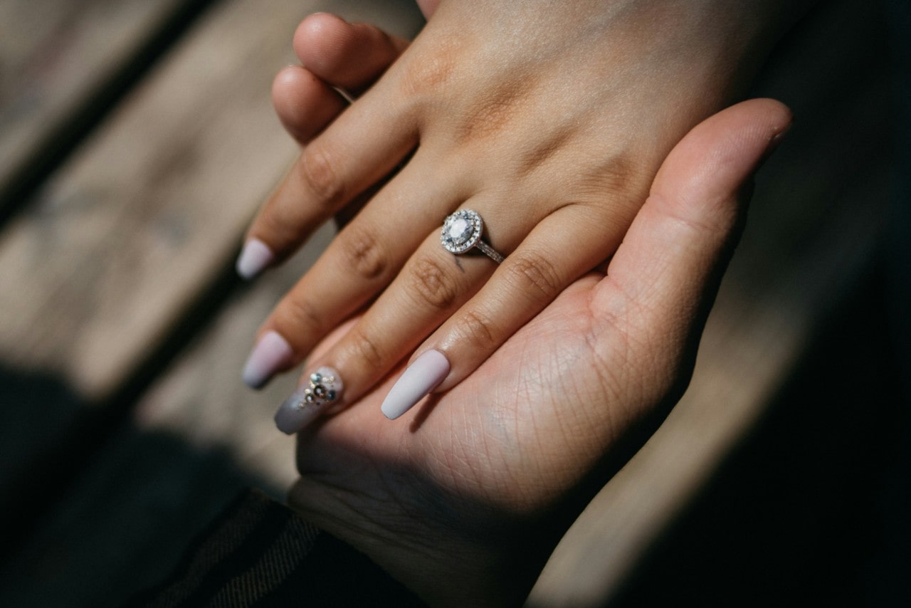 A woman’s hand with long nails showcase white gold halo diamond engagement ring, while being on top of man’s hand A woman’s hand with long nails showcase white gold halo diamond engagement ring, while being on top of man’s hand