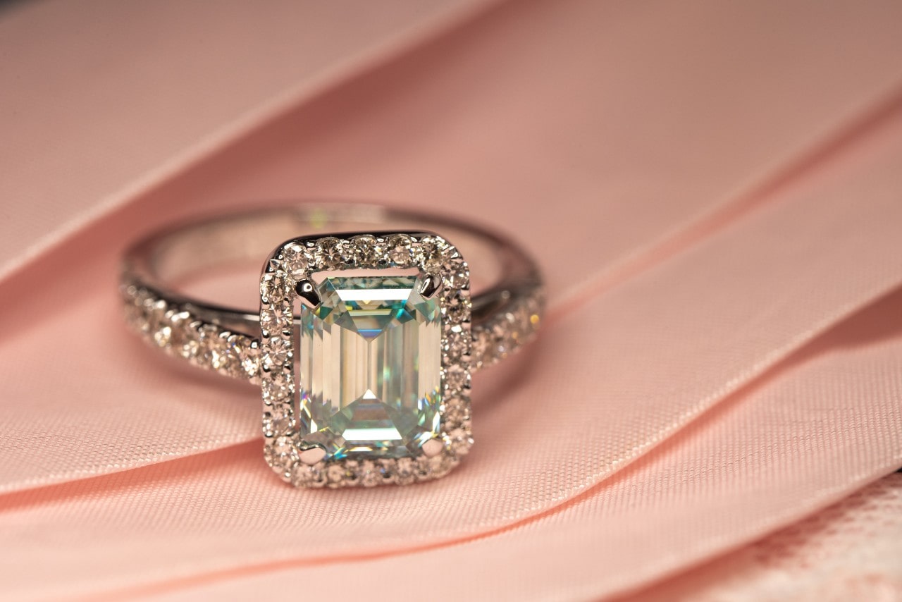 A close-up of an emerald-cut diamond ring with a halo setting and diamond-encrusted band, resting on pink fabric. A close-up of an emerald-cut diamond ring with a halo setting and diamond-encrusted band, resting on pink fabric.