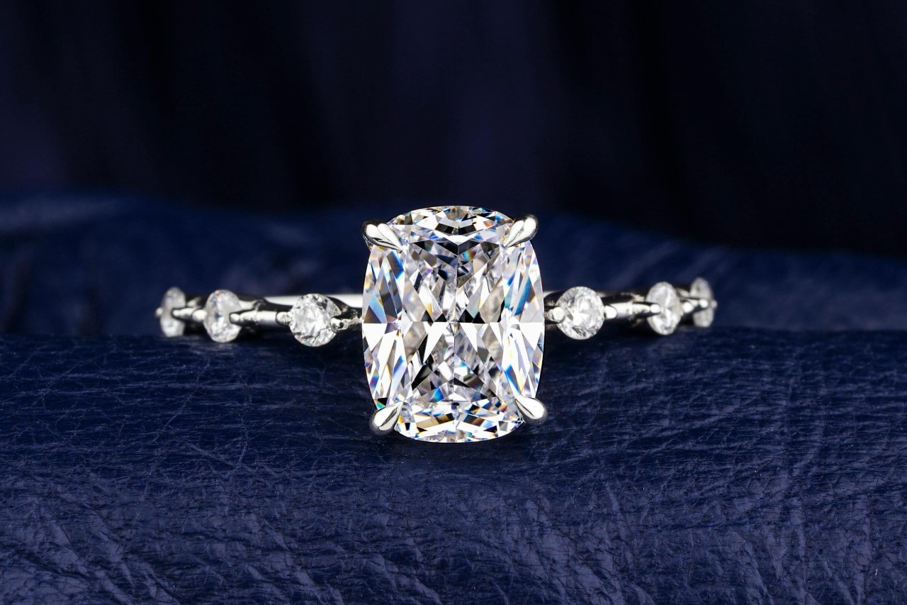 A close up of cushion cut white gold side stone engagement ring on a textured dark blue surface A close up of cushion cut white gold side stone engagement ring on a textured dark blue surface