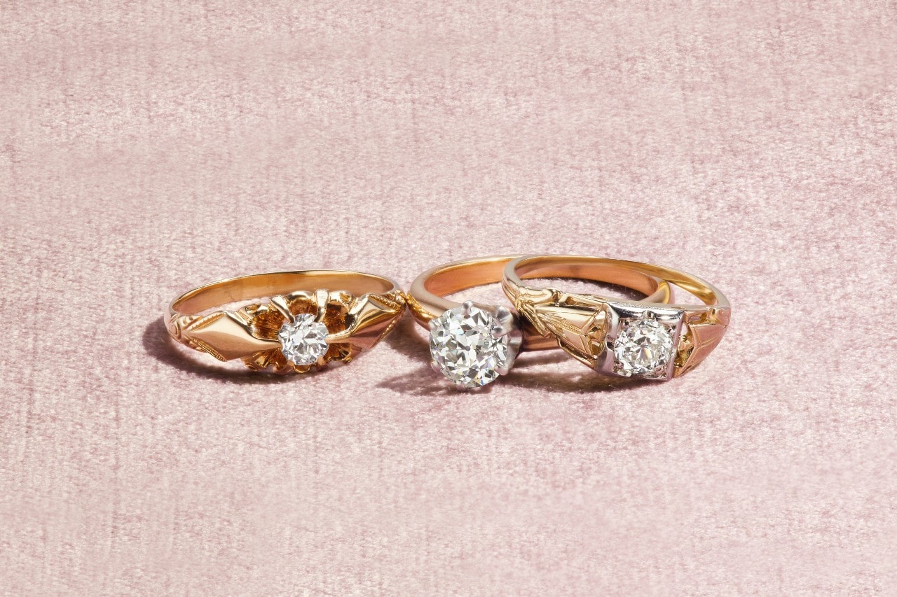 Three vintage gold rings with intricate designs and sparkling diamond centers are displayed on a soft pink textured surface.