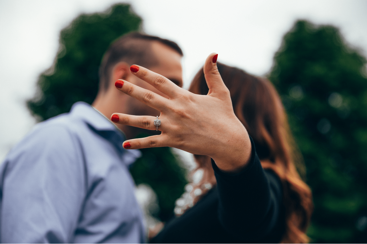 Close-up of an outstretched hand with a diamond engagement ring, foregrounding a couple sharing a romantic kiss outdoors.