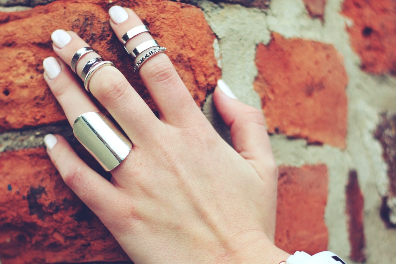 A woman’s hand sports several white gold fashion rings resting on a brick wall.