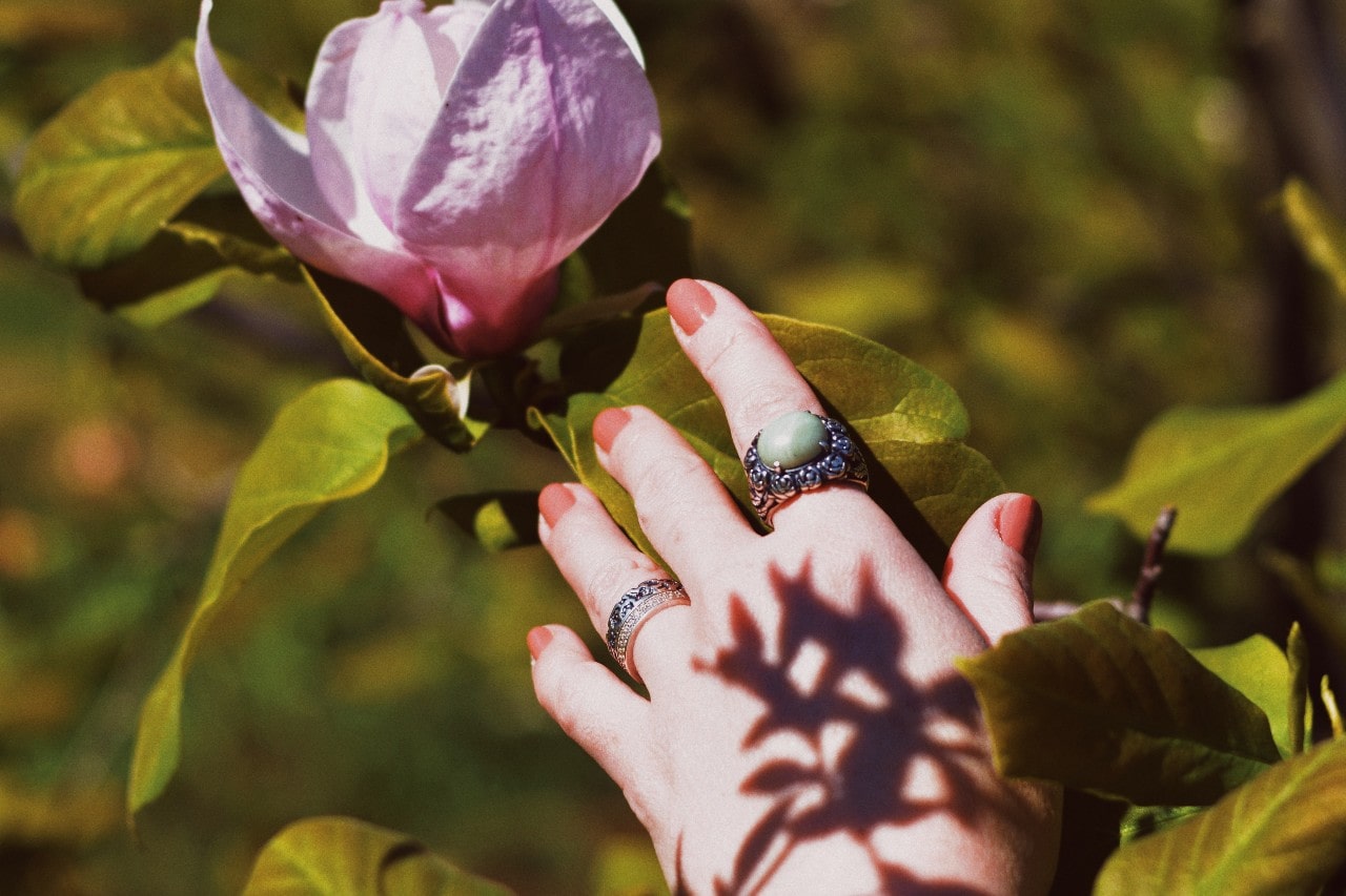 A woman wearing a gemstone fashion ring and two sterling silver rings reaches toward a large pink flower.
