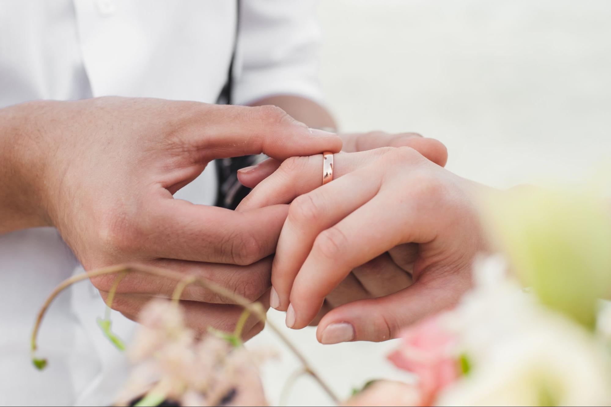 A close up of a hand placing a rose gold polished weeding band onto another hand. A close up of a hand placing a rose gold polished weeding band onto another hand.