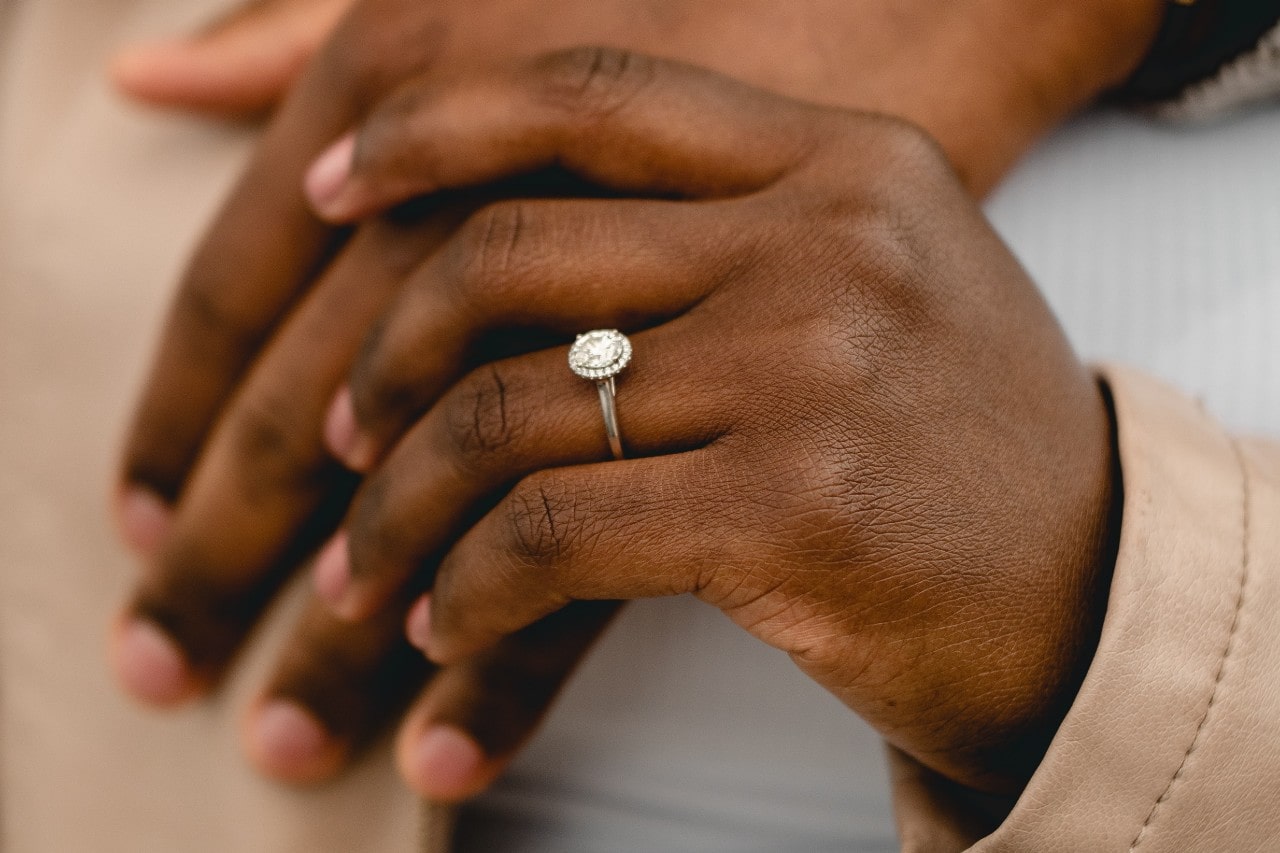 A hand in a pink leather jacket showcases white gold diamond oval cut halo engagement ring rests gently on another hand.