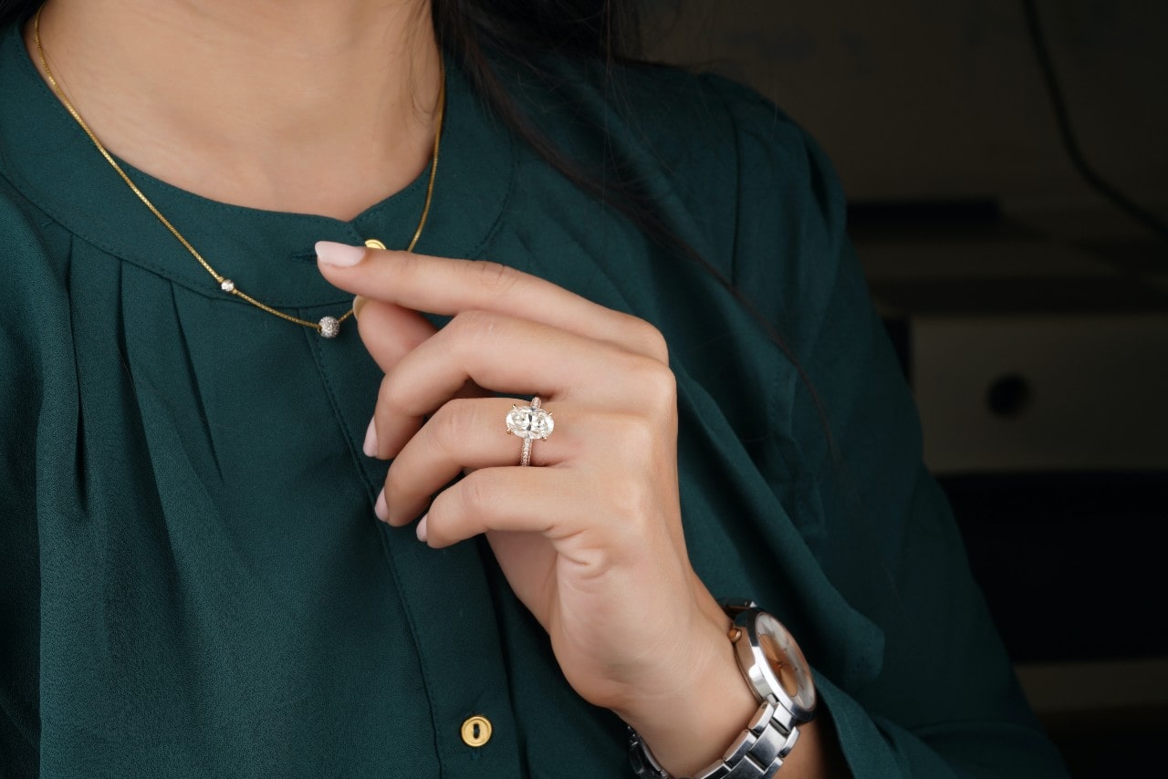 A woman in a green blouse displays her hand with an oval cut diamond engagement ring, while holding her necklace.