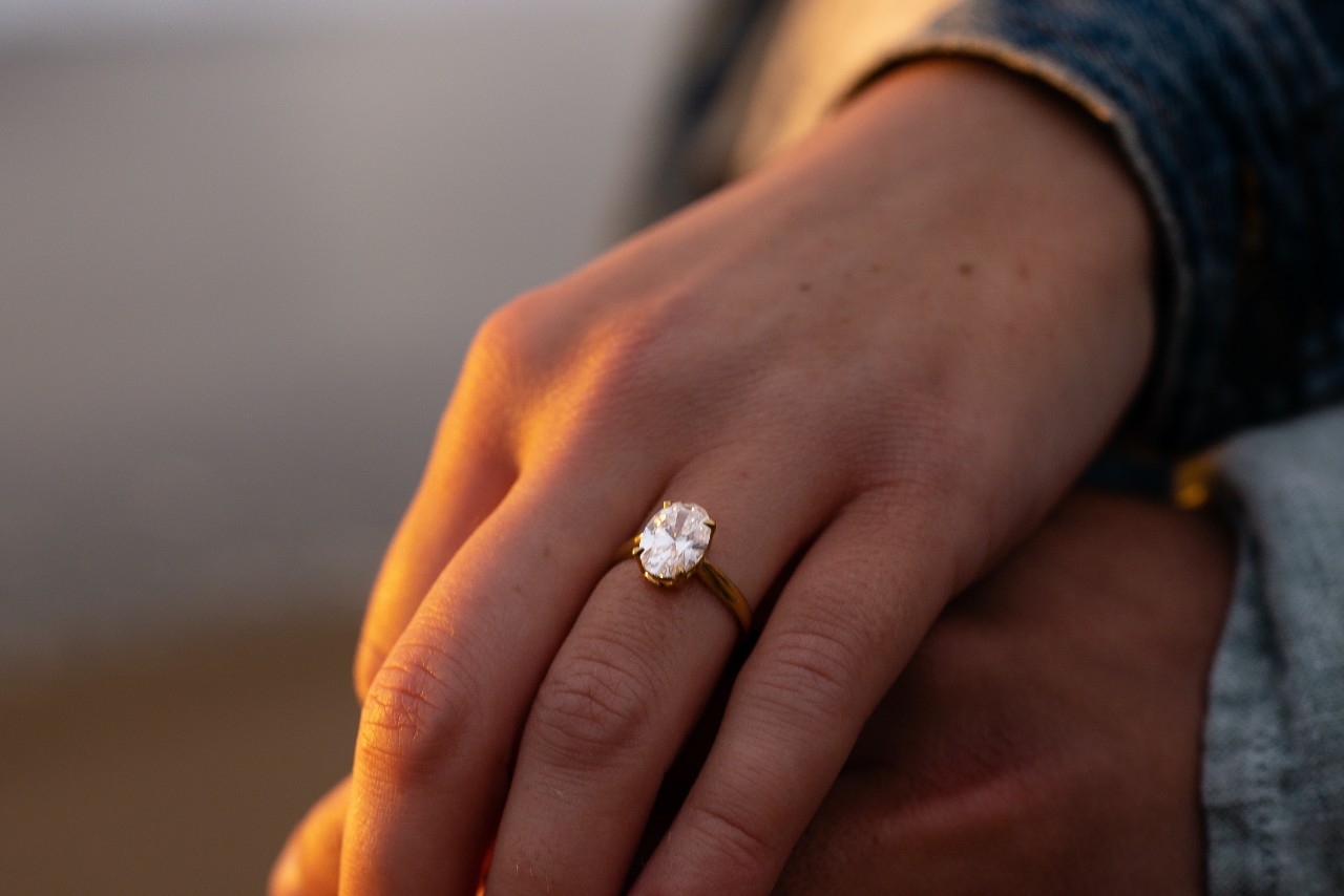 A hand wearing a yellow gold engagement ring with a large oval-cut diamond is shown in warm sunset light.