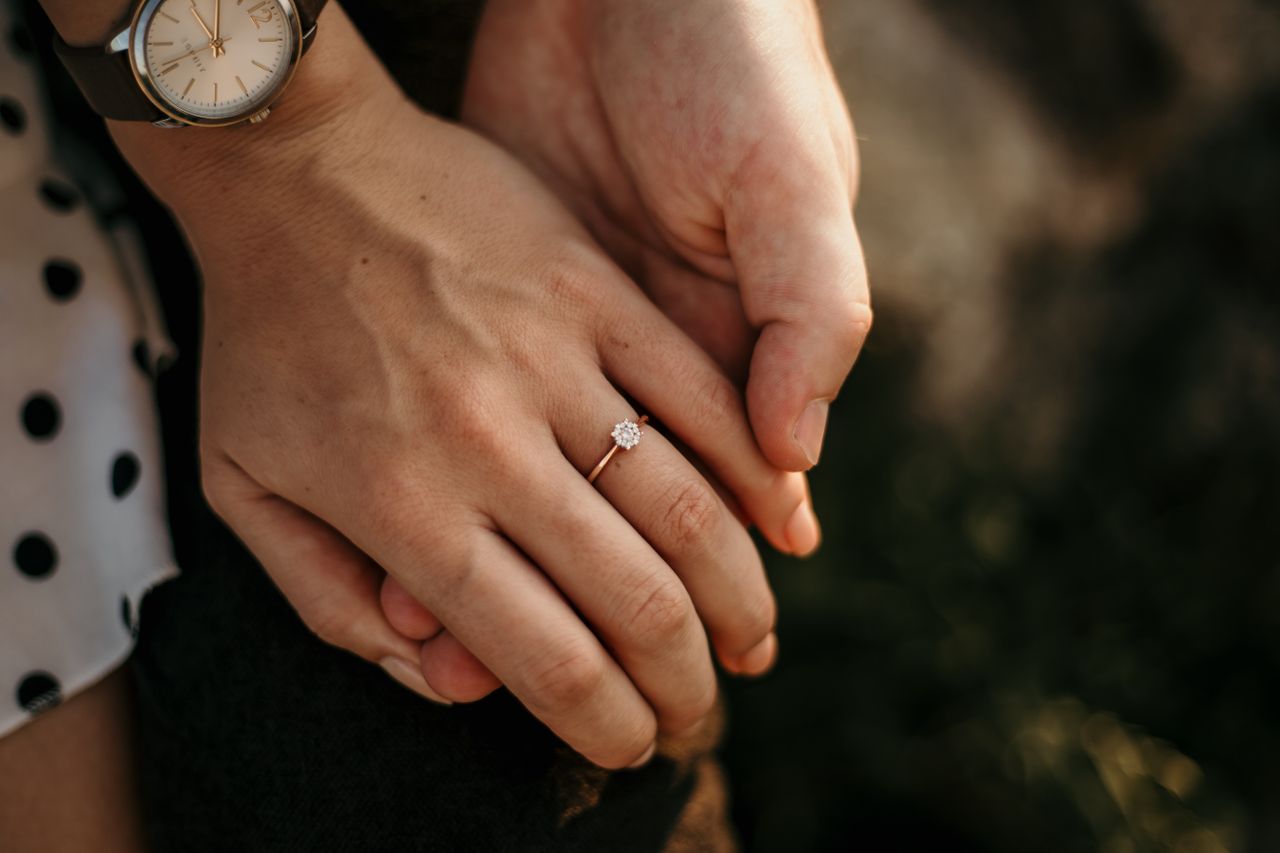 A man holds his fiancee’s hand, which sports a gold solitaire engagement ring.