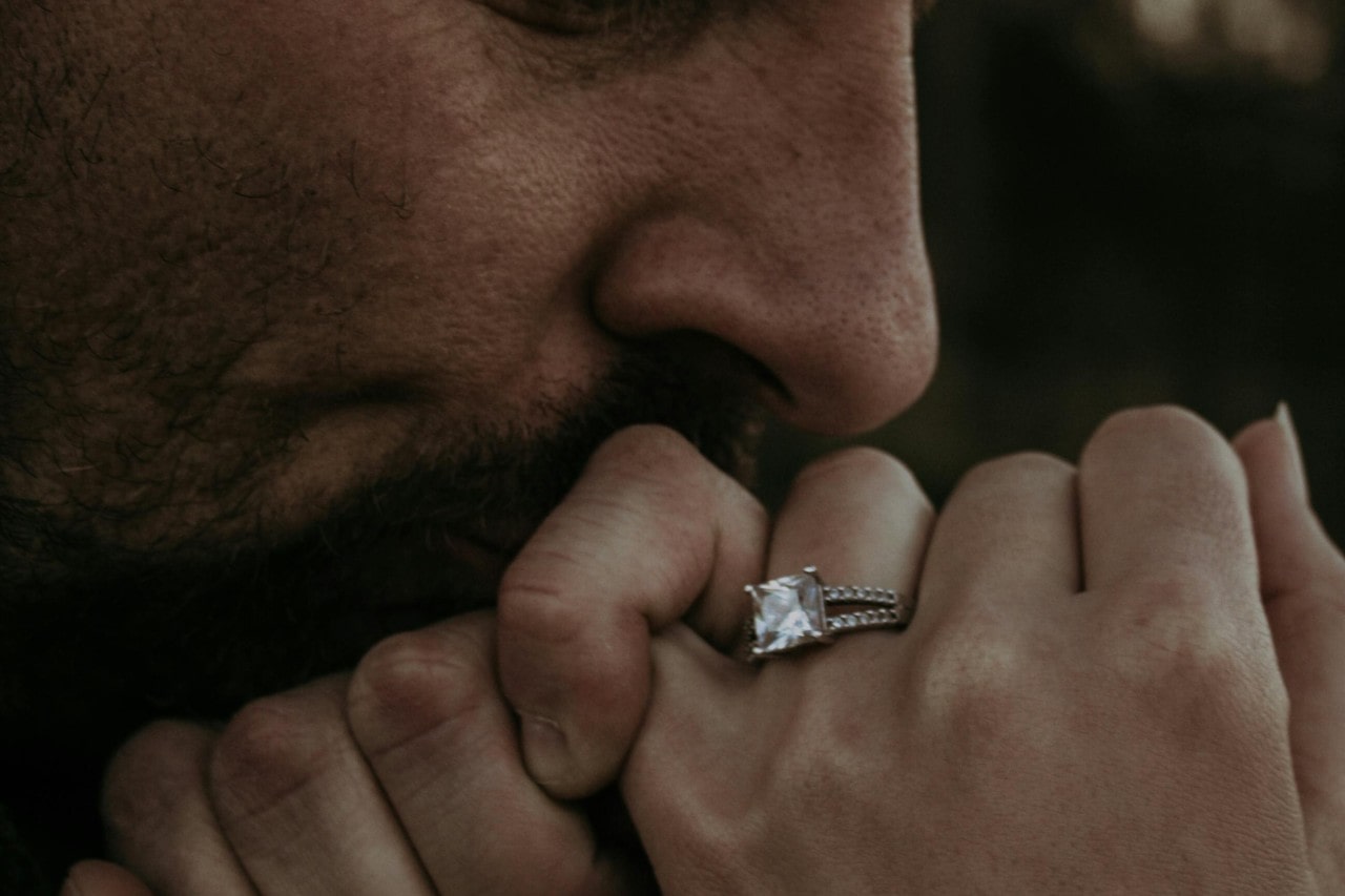 A man kissing a woman’s hand that is adorned with a princess cut engagement ring. A man kissing a woman’s hand that is adorned with a princess cut engagement ring.