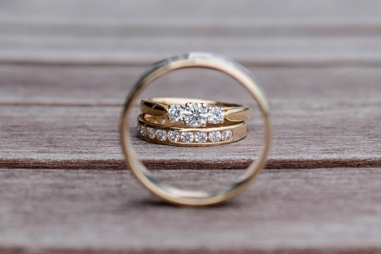 A pair of gold and diamond wedding rings is framed through another gold band, resting on a rustic wooden surface. A pair of gold and diamond wedding rings is framed through another gold band, resting on a rustic wooden surface.