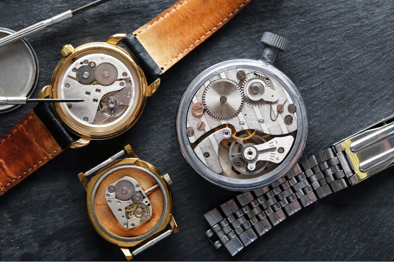 An assortment of vintage wristwatches with their cases open reveals mechanical movements and gears, accompanied by watchmaking tools.