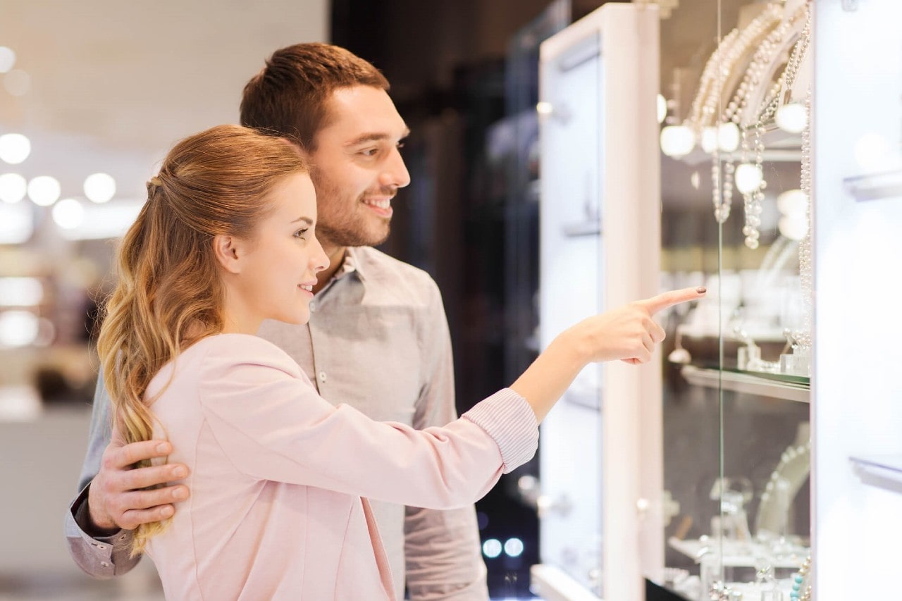 Smiling couple browsing store window; woman points at a display while her partner stands behind her, hand gently resting on her back.